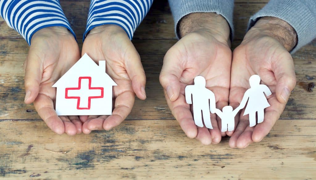 Hände halten Papierfamilie und Papierhaus mit roten Kreuz