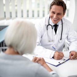 Positive emotional general practitioner sitting with the clipboard on the table and kindly smiling to his patient